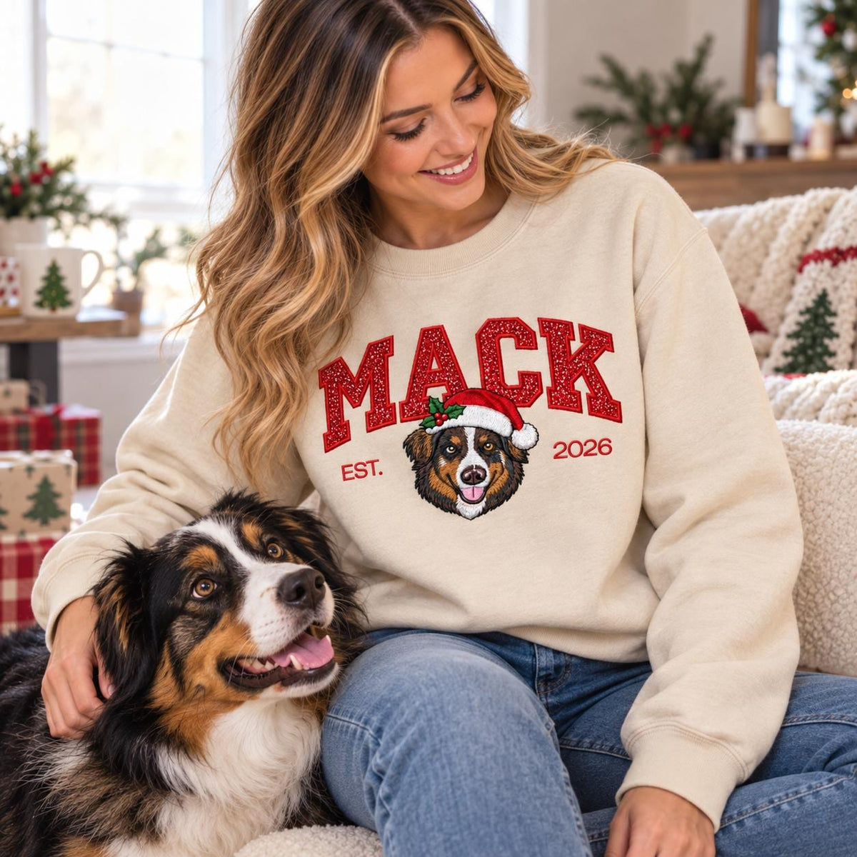 A woman sitting by a cozy fireplace wearing a customized Christmas dog sweatshirt in Sand color, featuring glitter text and an embroidered Australian Shepherd portrait, lovingly stroking her happy pet dog.