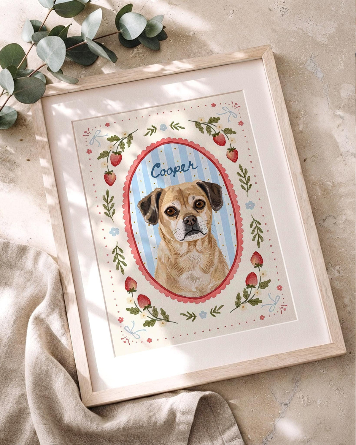 A framed illustration of a tan dog with dark ears, centered in a blue and white striped oval frame with a red scalloped border. The name Cooper is written above the dog. The border features red strawberries, green leaves, and blue bows.