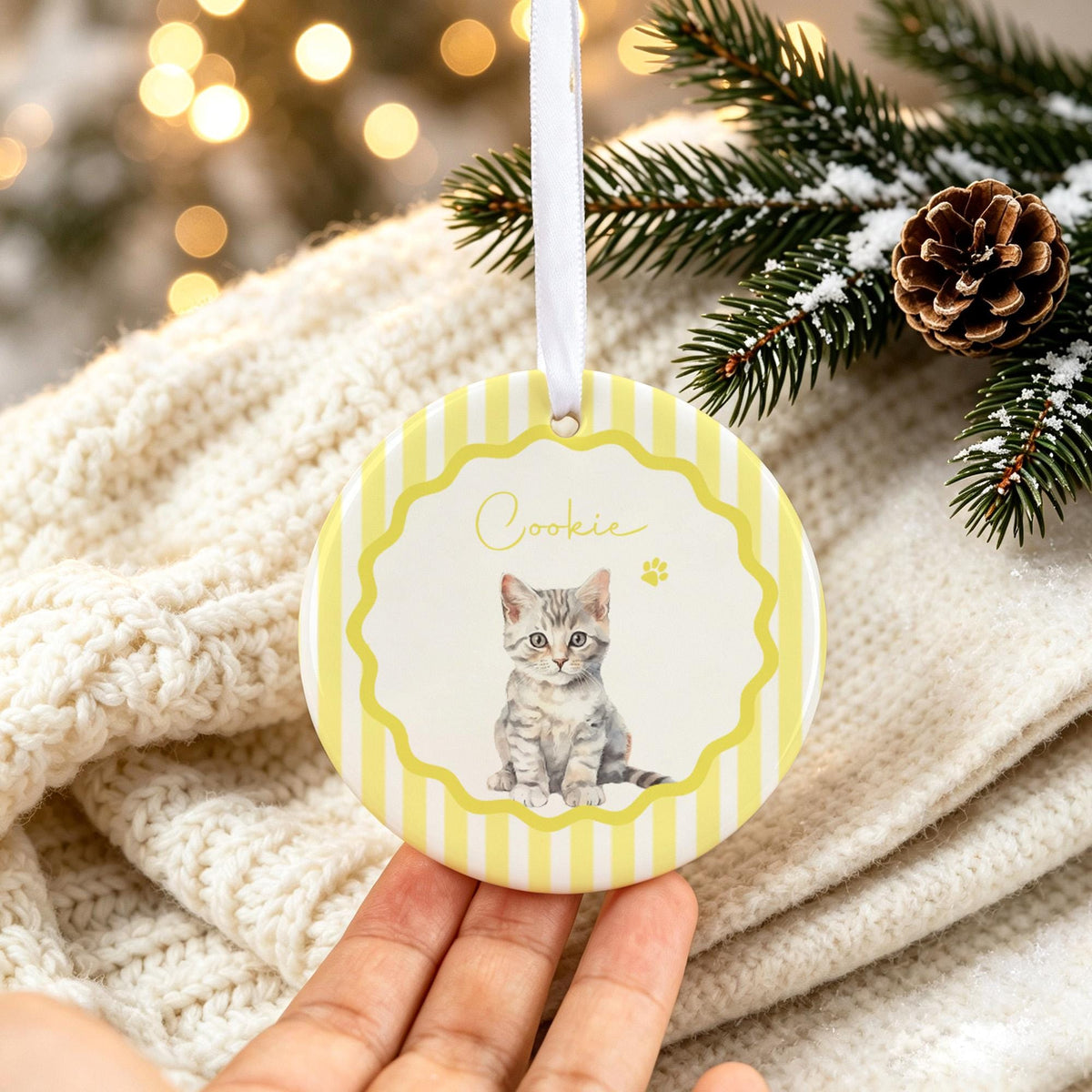 A round ceramic ornament featuring a white center with a gray tabby kitten illustration and the name Cookie written in cursive. The border is yellow with white vertical stripes and a scalloped edge, suspended by a white ribbon.