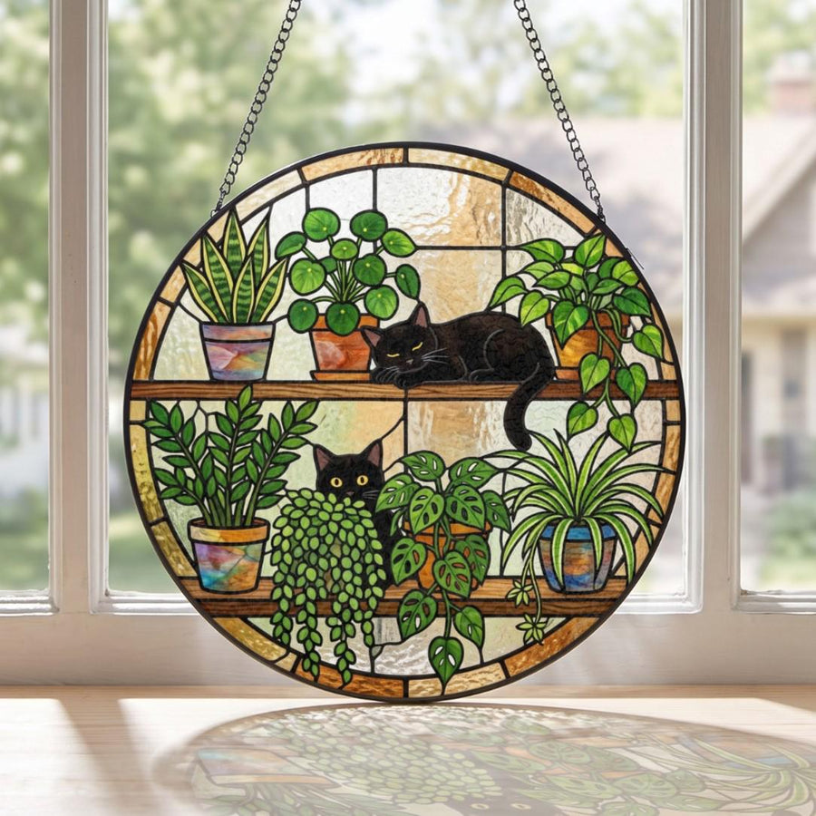 A circular stained glass suncatcher featuring two black cats resting on wooden shelves among various potted houseplants. The design includes a snake plant, a pilea, a trailing vine, and a spider plant, all rendered in vibrant glass.