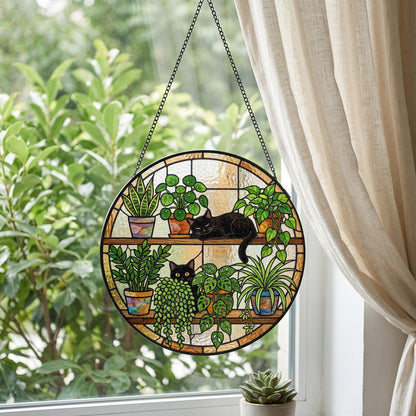 A circular stained glass suncatcher featuring two black cats resting on shelves among various potted houseplants. The design includes a snake plant, a pilea, a trailing pothos, and other green foliage in colorful pots, suspended by a black chain.