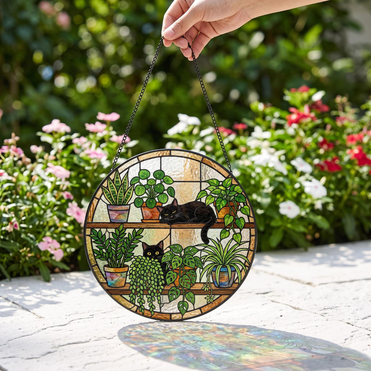 A circular stained glass suncatcher featuring two black cats resting among various potted plants on wooden shelves. The design includes green foliage, colorful pots, and a black metal chain for hanging, creating a detailed scene.
