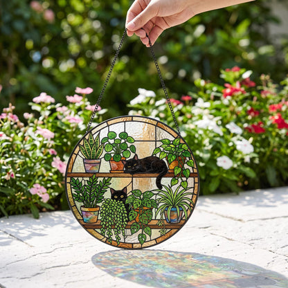 A circular stained glass suncatcher featuring two black cats resting among various potted plants on wooden shelves. The design includes green foliage, colorful pots, and a black metal chain for hanging, creating a detailed scene.