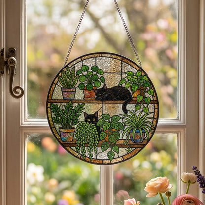 A circular stained glass suncatcher featuring two black cats resting among various potted green plants on two shelves. The piece is framed in a dark metal border and hangs from a silver-colored chain against a window pane.