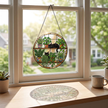 A circular stained glass suncatcher featuring two black cats resting among various potted green plants on two shelves. The piece is suspended by a metal chain and displays colorful glass segments in shades of green, brown, and amber.