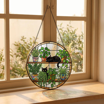 A circular stained glass suncatcher featuring two black cats resting among various potted green plants on two shelves. The piece is framed in dark metal and hangs from a thin metal chain, displaying vibrant colors and textures.