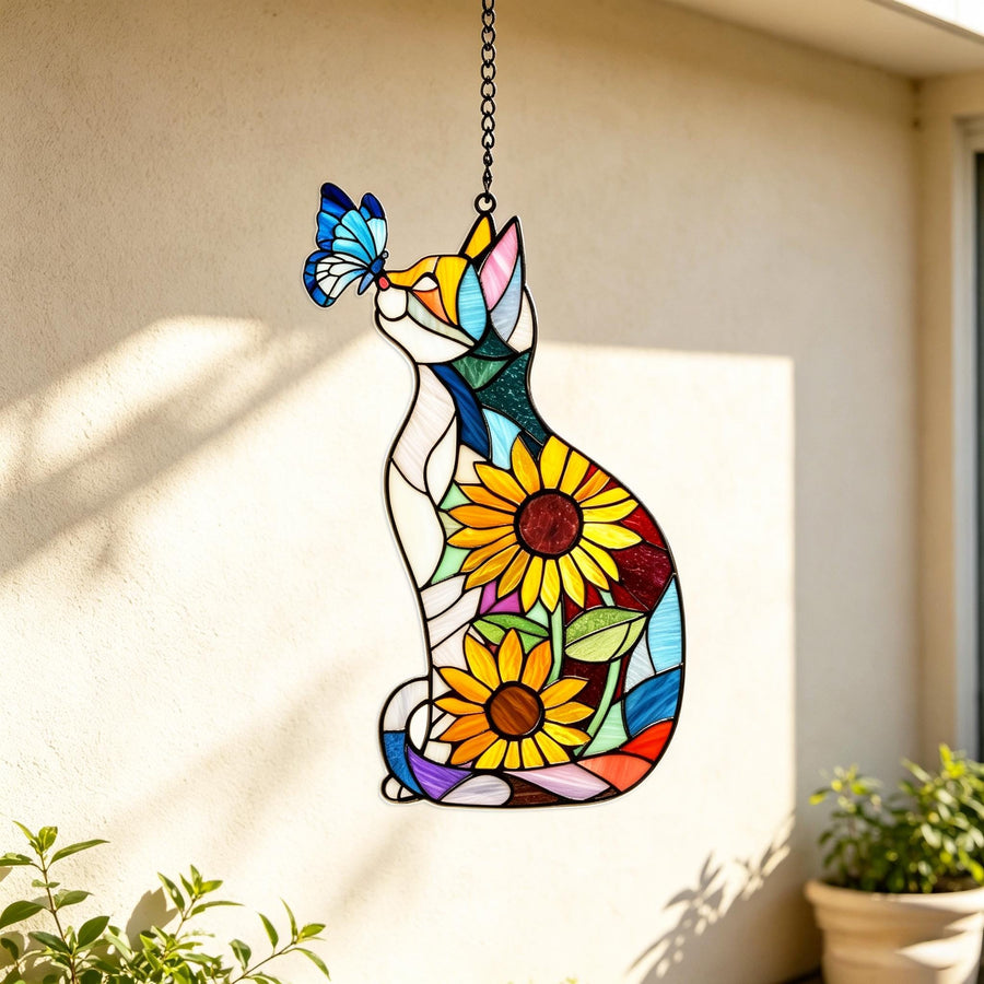 A stained glass suncatcher shaped like a sitting cat with a blue butterfly perched on its nose. The cat features a colorful mosaic pattern including two large yellow sunflowers with brown centers, green leaves, and various blue and white glass pieces.