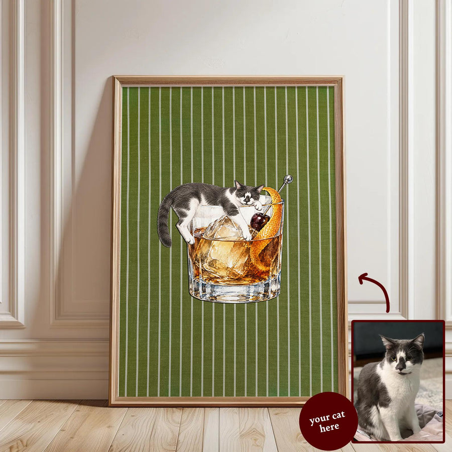 A framed wall art print featuring a grey and white cat lounging inside a glass of amber-colored cocktail with ice, a cherry, and an orange peel. The background is a solid olive green with thin vertical white stripes.