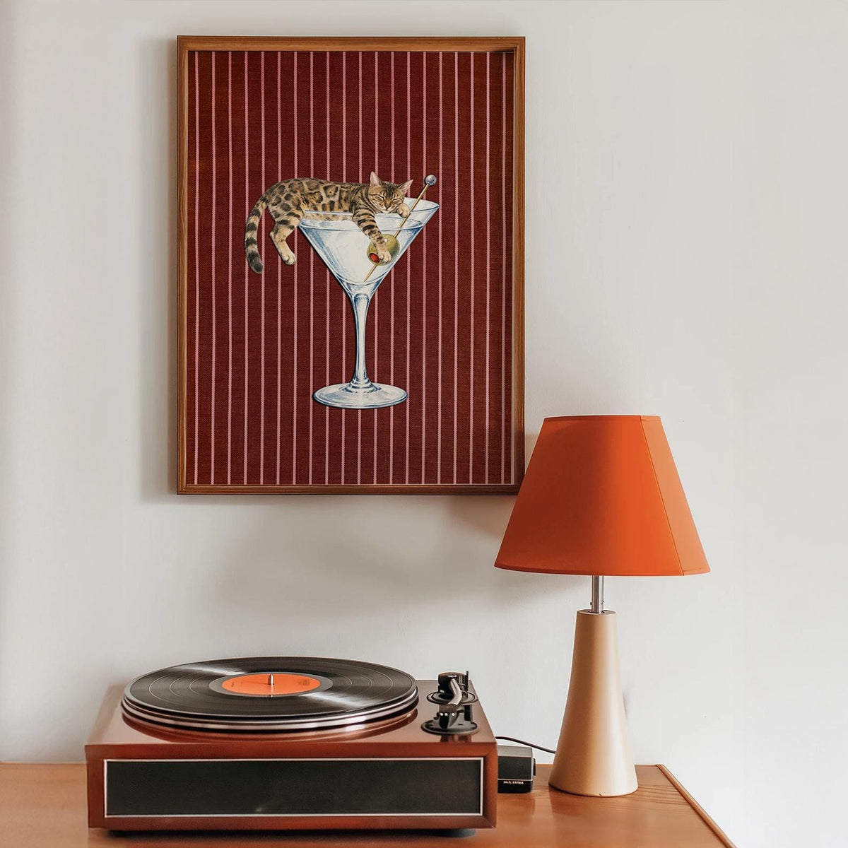 A framed wall art print featuring a brown tabby cat lounging inside a large glass martini glass with a single green olive on a pick. The background of the print is a deep red with thin vertical white pinstripes and a wood frame.