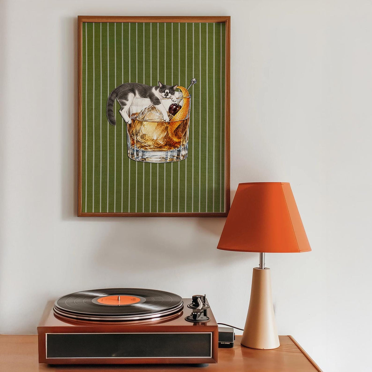 A framed wall art print featuring a black and white cat lounging inside a glass of amber-colored cocktail with ice, a cherry, and an orange peel garnish, set against a dark green background with thin vertical white stripes.