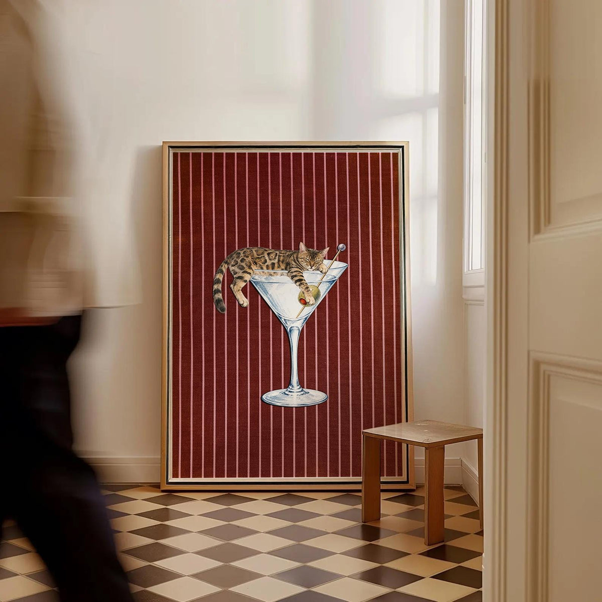 A rectangular framed wall art print featuring a brown spotted cat lounging inside a large martini glass with an olive on a toothpick. The background consists of a deep red fabric texture with vertical white pinstripes throughout.