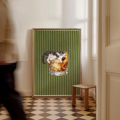 A framed rectangular wall art piece featuring a green background with thin white vertical stripes. The central illustration depicts a glass of amber-colored liquid with ice, a dark cherry, and an orange peel, topped by a black and white cat.