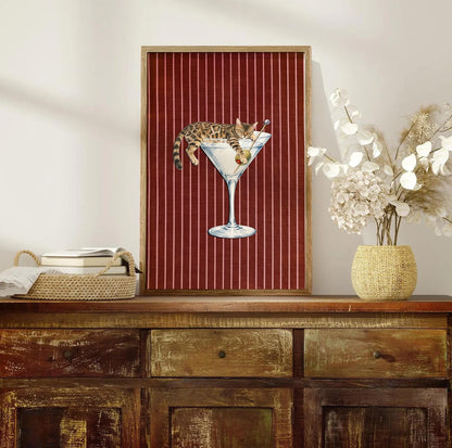 A rectangular wall art print in a wooden frame featuring a brown tabby cat lounging inside a large martini glass with an olive on a toothpick. The background is a deep red fabric texture with thin vertical white pinstripes.