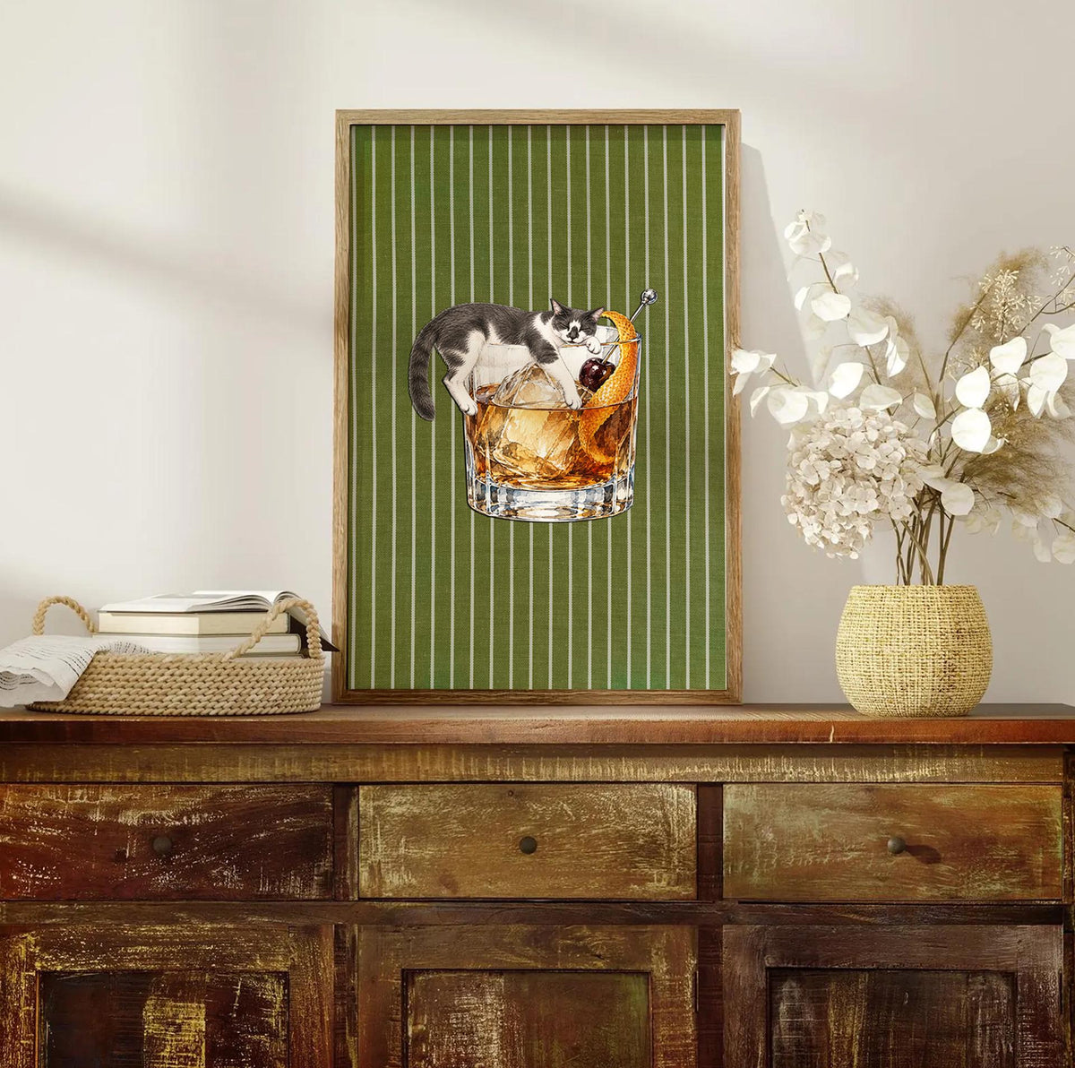 A framed vertical wall art print featuring a green background with thin white vertical stripes. Centered is a graphic of a black and white cat lounging on top of a glass of amber-colored liquid with ice, an orange peel, and a cherry.