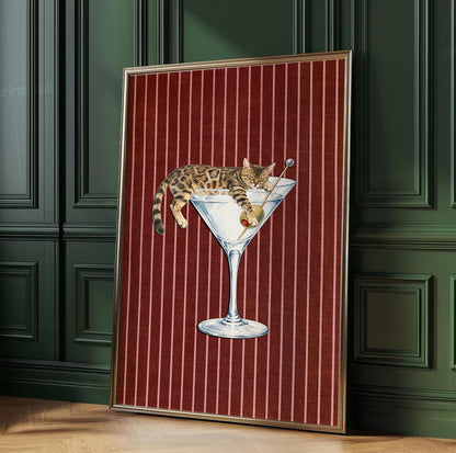 A framed wall art print featuring a brown tabby cat resting inside a large glass martini glass. The cat is draped over the rim with a green olive on a toothpick. The background is a deep red fabric texture with vertical white stripes.