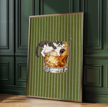 A framed wall art print featuring a grey and white cat lounging over a glass of amber-colored liquid with ice cubes, a dark red cherry, and an orange peel garnish, set against a vertical olive green background with thin white stripes.