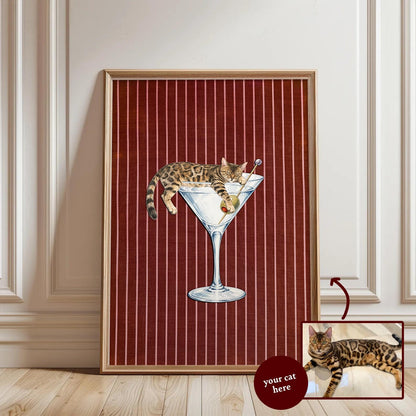 A framed vertical wall art print featuring a brown spotted tabby cat lounging inside a large martini glass with an olive on a toothpick. The background is a deep red fabric texture with thin vertical white pinstripes throughout.