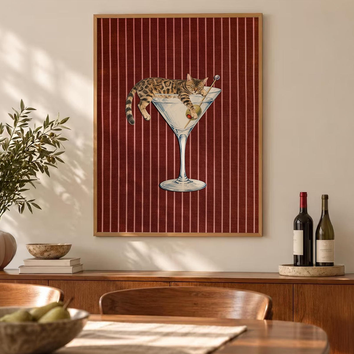 A framed rectangular wall art print featuring a brown tabby cat lounging inside a large glass martini glass. The cat is positioned with its paws draped over the rim, next to a green olive on a pick. The background is a deep red with thin white vertical stripes.