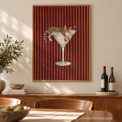 A framed rectangular wall art print featuring a brown tabby cat lounging inside a large glass martini glass. The cat is positioned with its paws draped over the rim, next to a green olive on a pick. The background is a deep red with thin white vertical stripes.