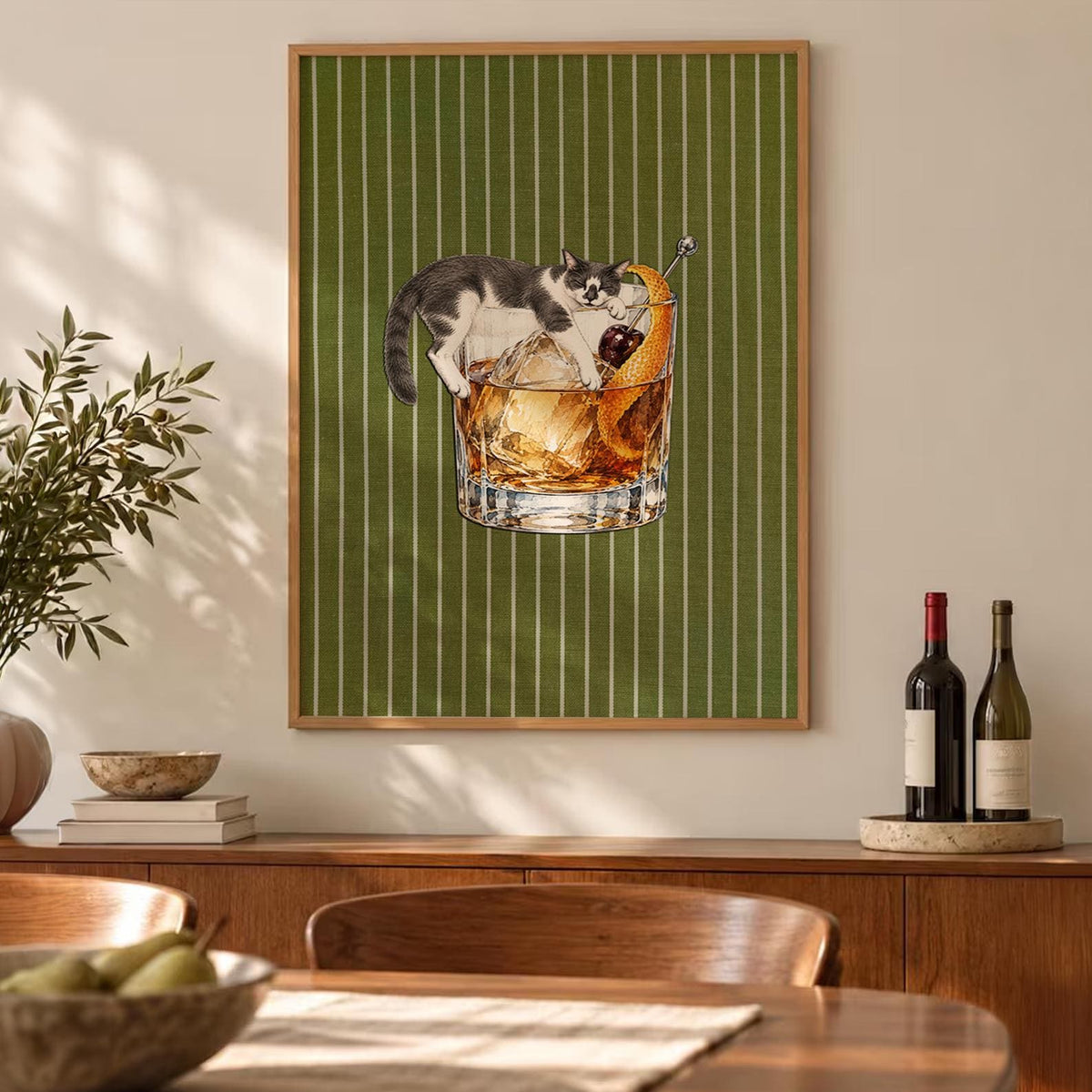 A framed wall art print featuring a grey and white cat lounging inside a glass of amber-colored cocktail with a large ice cube, a dark cherry, and an orange peel garnish, set against a vertical white-striped olive green background.
