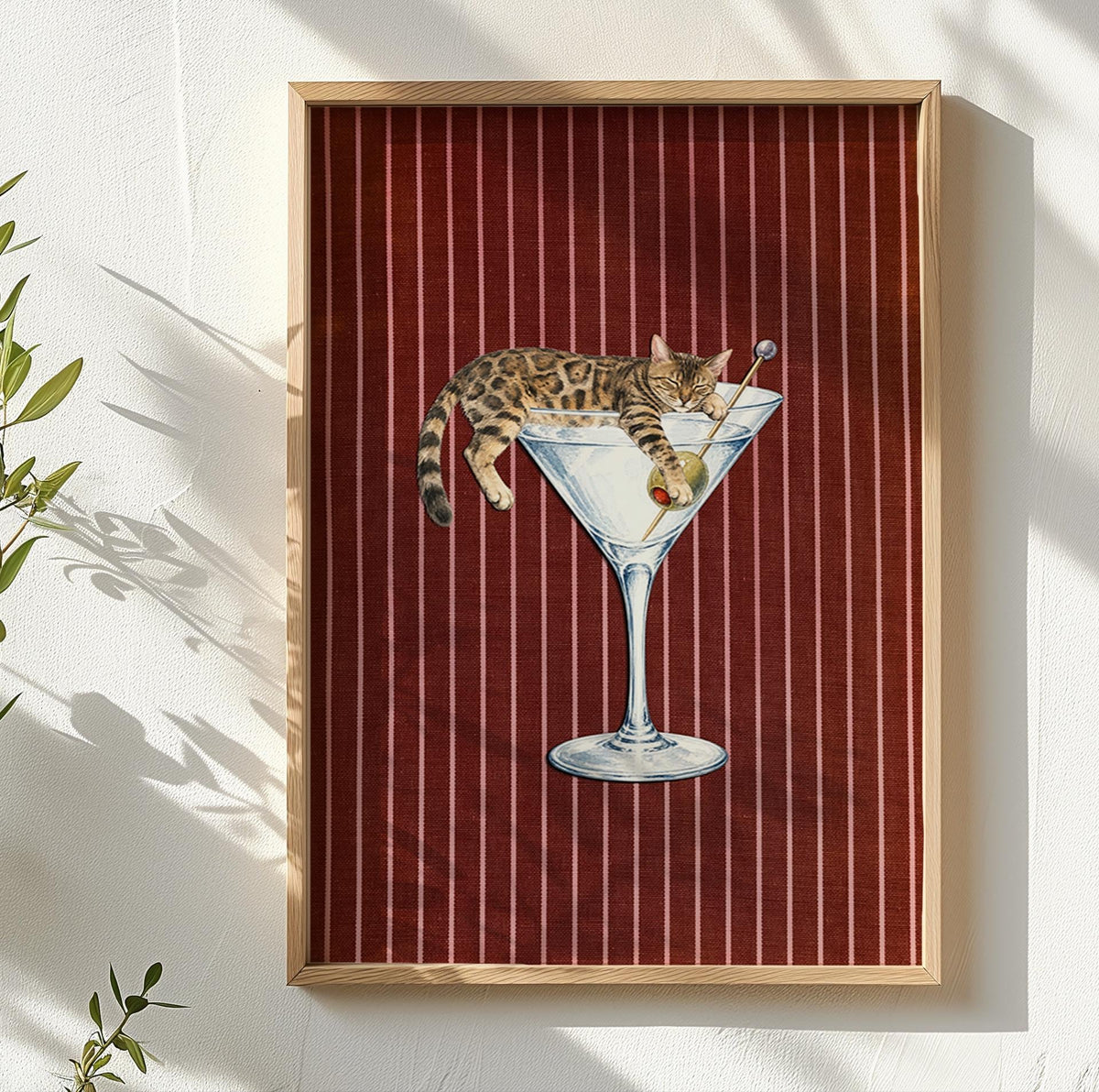 A framed wall art print featuring a brown tabby cat lounging inside a large martini glass. The cat holds a toothpick with a green olive. The background consists of a deep red fabric texture with thin, vertical white pinstripes.