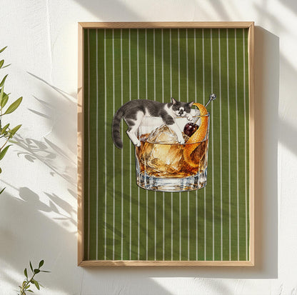 A framed wall art print featuring a black and white cat lounging inside a glass of amber-colored liquid with ice cubes, a dark cherry, and an orange peel garnish, set against a vertical white-striped olive green background.