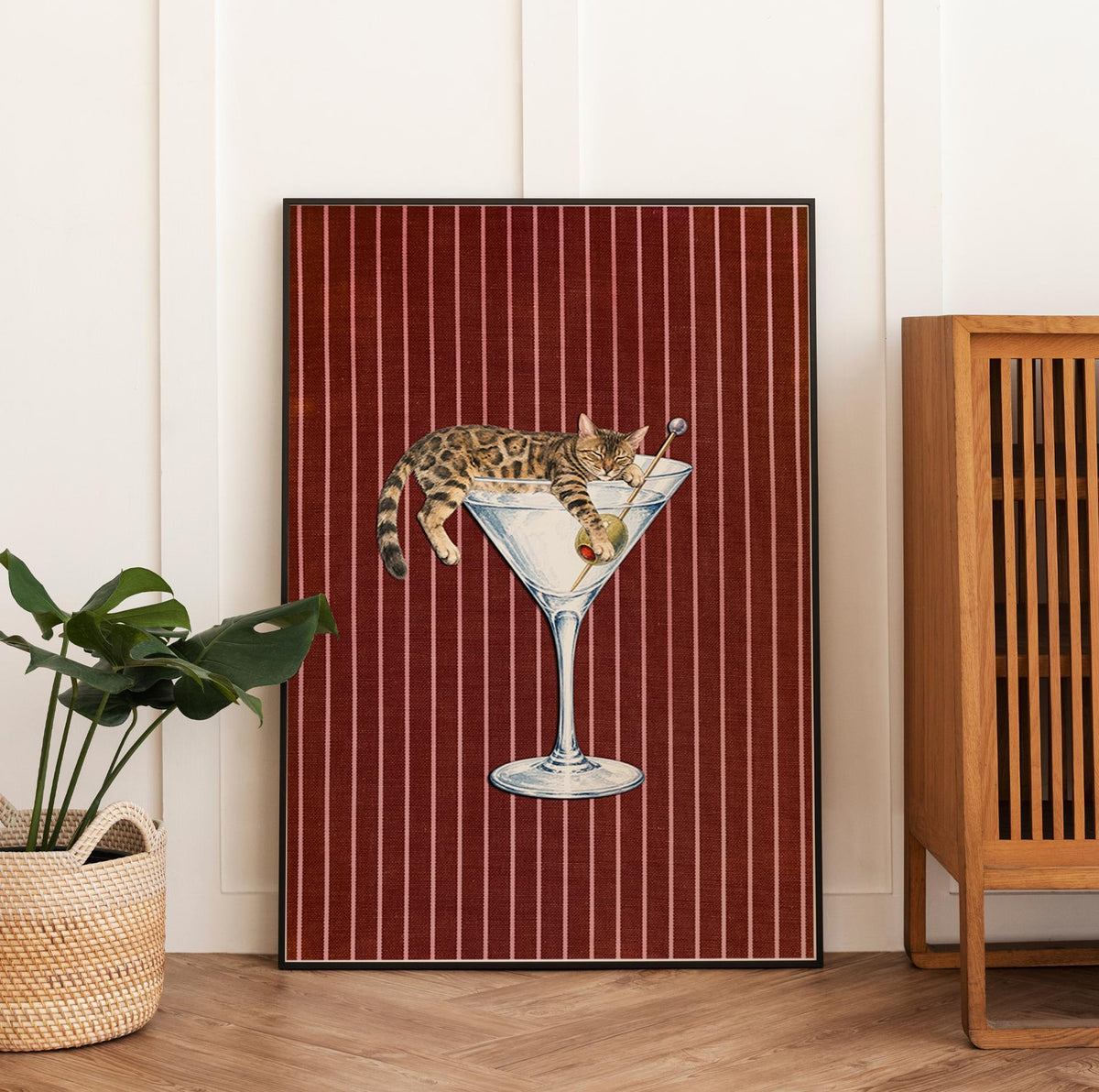 A rectangular wall art print featuring a brown tabby cat resting inside a large martini glass. The cat holds a green olive on a toothpick. The background consists of a deep red fabric texture with thin, vertical white stripes.