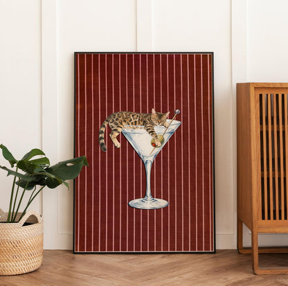 A rectangular wall art print featuring a brown tabby cat resting inside a large martini glass. The cat holds a green olive on a toothpick. The background consists of a deep red fabric texture with thin, vertical white stripes.