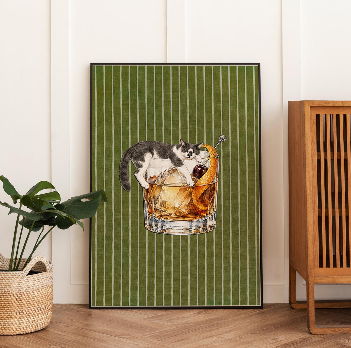 A framed wall art print featuring a black and white cat lounging over a glass of amber-colored cocktail with ice cubes, a dark cherry, and an orange peel garnish, set against a vertical olive green background with thin white stripes.