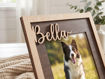 A wooden picture frame featuring a light-colored outer border and a dark-colored inner panel. The name Bella is cut out in a light-colored script font and attached to the top of the frame. A photograph of a black and white dog is displayed.