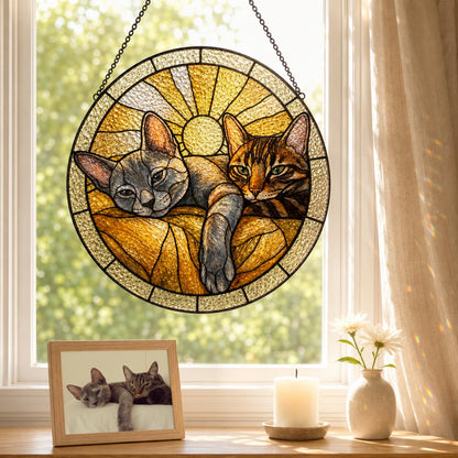 A circular stained glass suncatcher featuring two cats resting together against a golden sunburst background. The cats are rendered in shades of gray and brown, with one cat lying in front of the other. The piece is framed in black metal.