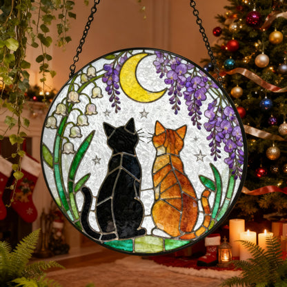 A round stained glass window featuring two cats gazing at a crescent moon. The cats are black and orange, set against a backdrop of purple wisteria, white flowers, and green foliage. The window is hanging from a chain.