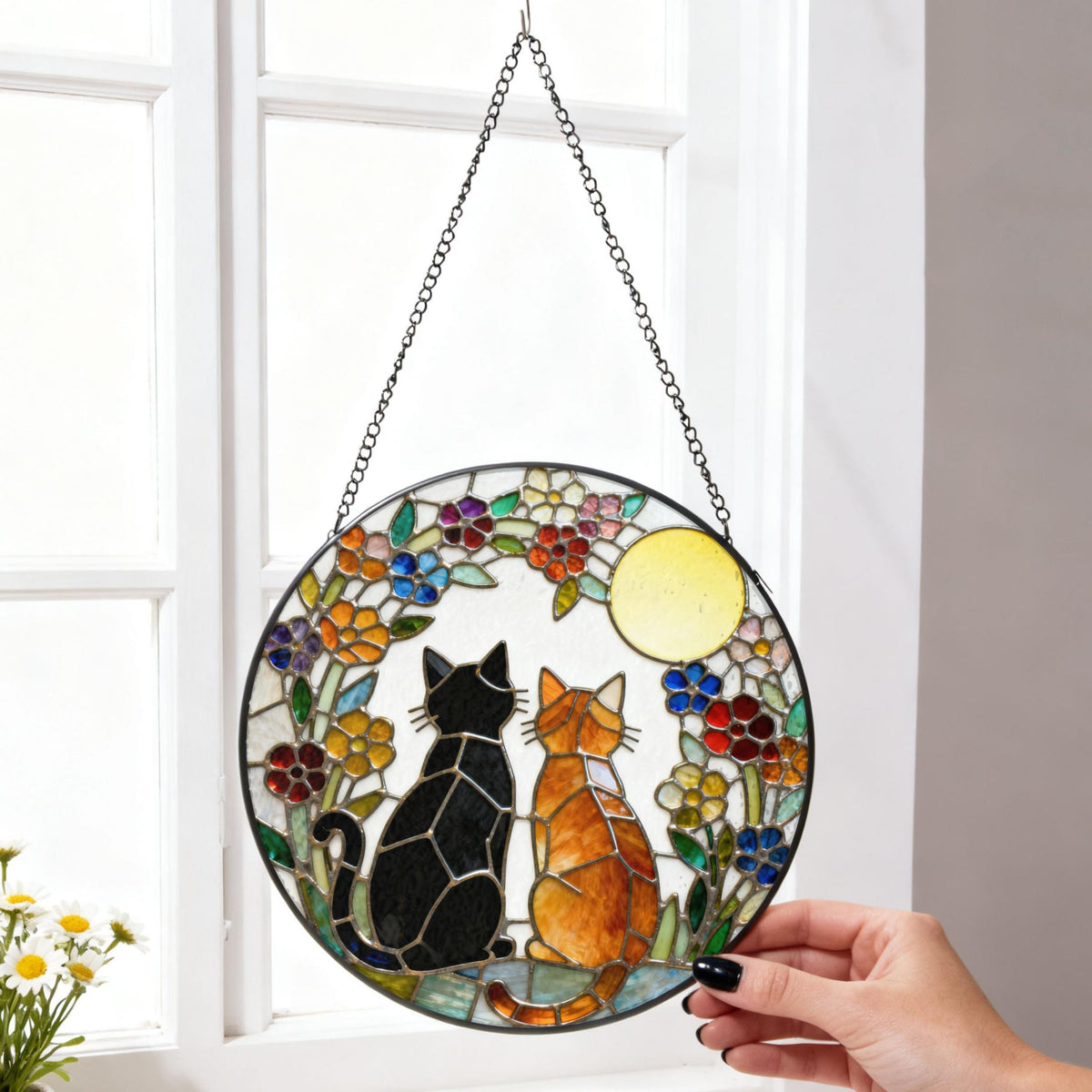 Stained glass round window hanging featuring two cats, one black and one orange, gazing at a yellow moon. The cats are surrounded by colorful flowers. The piece is suspended by a chain.