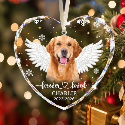 A heart-shaped clear acrylic ornament featuring a golden retriever with white angel wings, white snowflakes, and gold stars. The text reads forever loved, CHARLIE, and 2022-2026. It hangs from a white ribbon against a blurred holiday background.