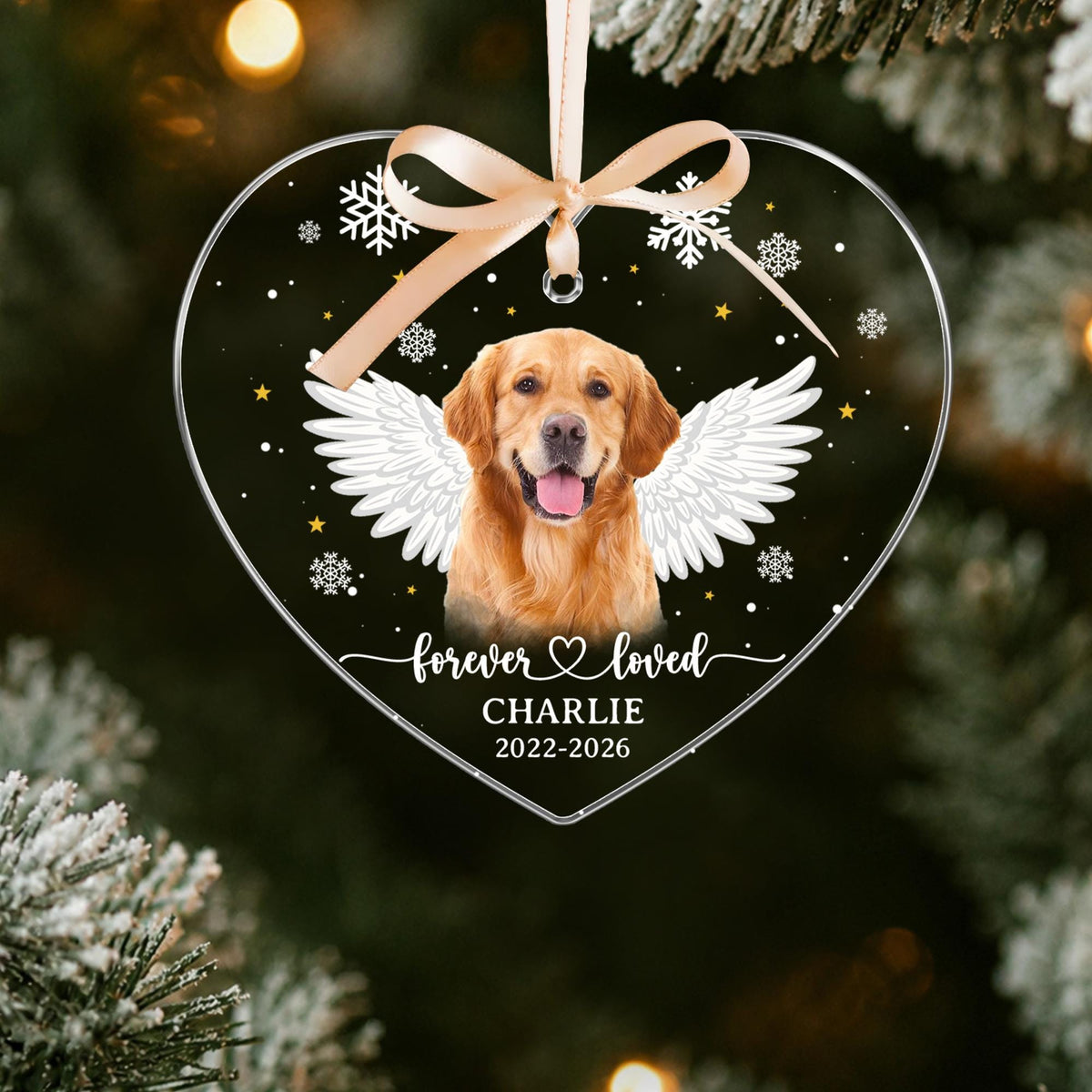 A heart-shaped clear acrylic ornament featuring a golden retriever with white angel wings. The design includes white snowflakes, gold stars, and the text "forever loved", "CHARLIE", and "2022-2026". A beige ribbon is tied at the top.