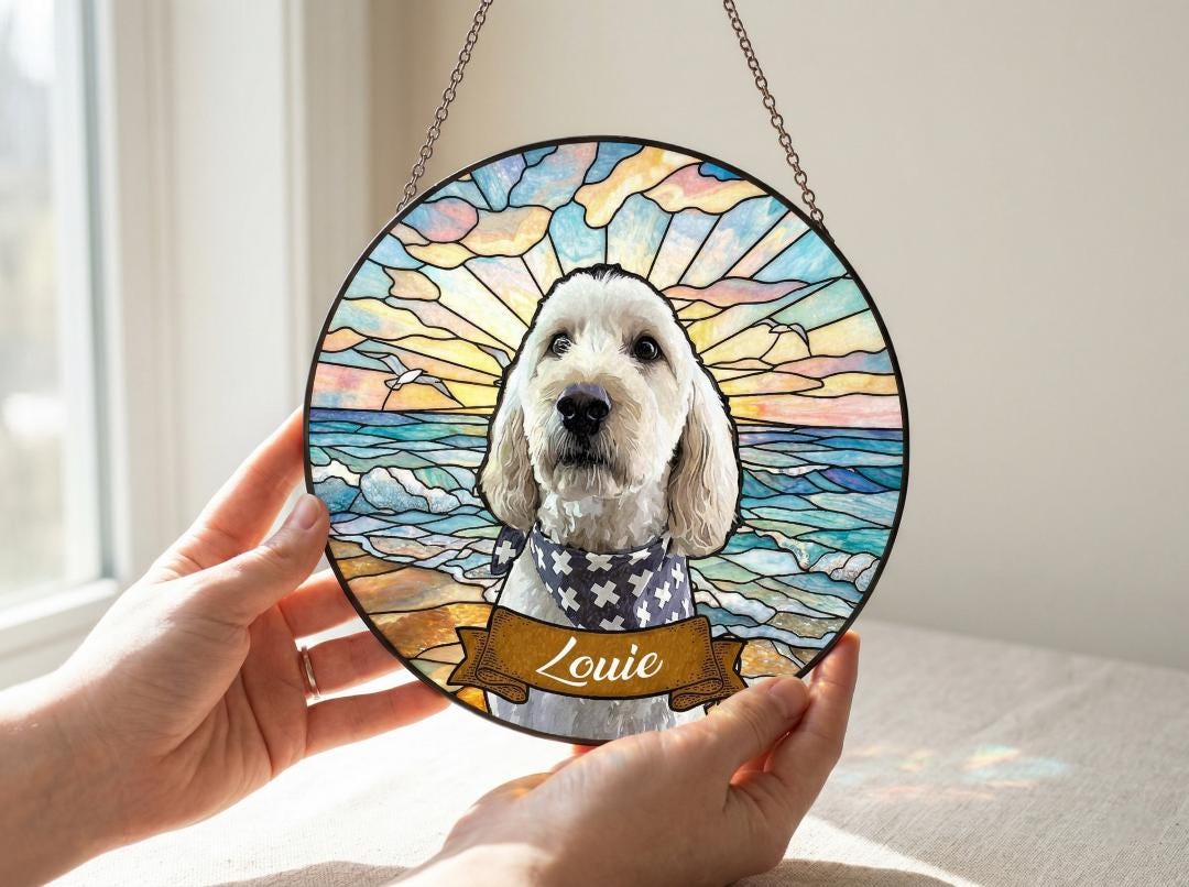 Round stained glass window art featuring a dog named Louie. The artwork depicts a dog with a blue and white bandana, set against a beach scene with a sunset and ocean waves. The piece is held by two hands.