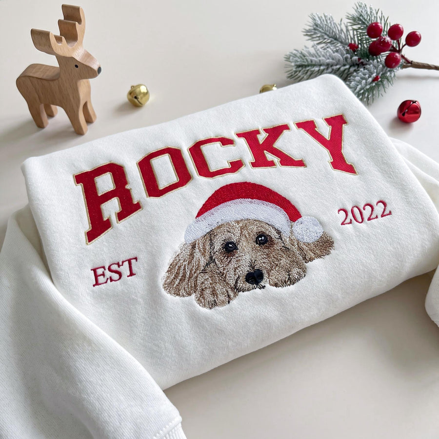 A white sweatshirt featuring red embroidered text that reads ROCKY, EST, and 2022. Below the name is an embroidered illustration of a light brown dog wearing a red and white Santa hat. The design is centered on the front of the garment.