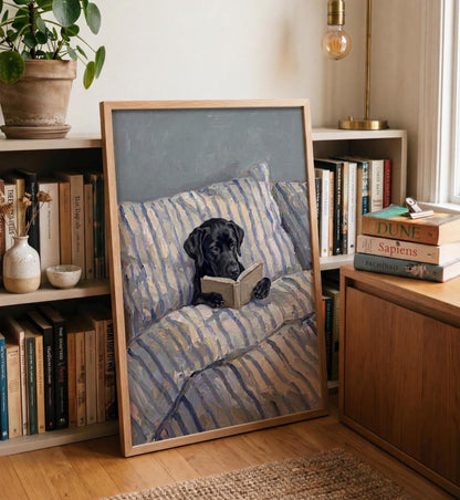 A framed rectangular wall art print featuring an oil painting of a black Labrador dog lying on a blue and white striped pillow while holding an open book. The artwork is displayed on a wooden shelf next to various books and a plant.