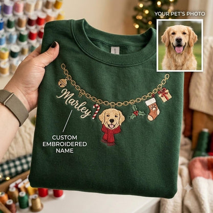 A dark green sweatshirt featuring an embroidered gold chain design with a bell, a candy cane, a golden retriever wearing a red scarf, holly, and a stocking. The name Marley is embroidered in white script, and a photo of a dog is shown.