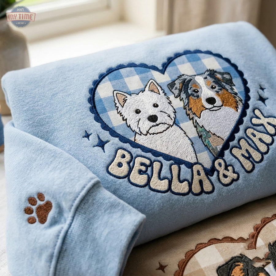 A light blue sweatshirt featuring an embroidered heart-shaped patch with a blue and white gingham background. The patch displays a white Westie and a tricolor Australian Shepherd, with the embroidered text Bella & Max below.