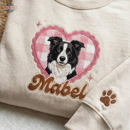 A beige crewneck sweatshirt featuring an embroidered design of a black and white Border Collie dog inside a pink gingham heart. Below the heart, the name Mabel is embroidered in brown script, and a brown paw print is on the sleeve.