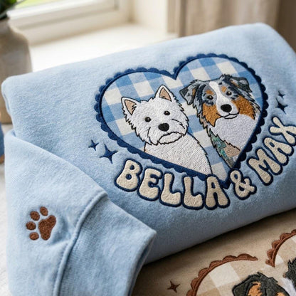 A light blue sweatshirt featuring an embroidered heart-shaped patch with a blue and white gingham pattern. Inside the heart are two dogs, a white Westie and a tricolor Australian Shepherd, with the text BELLA & MAX and a paw print.