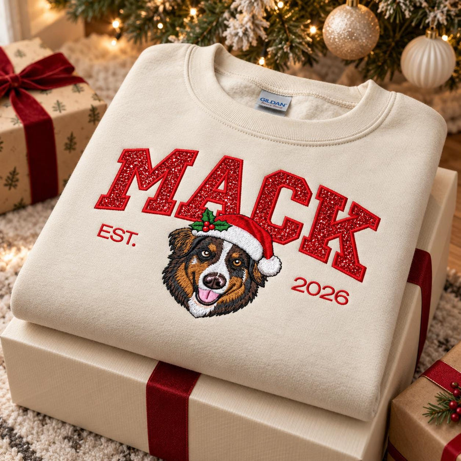 A cozy holiday scene featuring a folded Sand-colored custom Christmas dog sweatshirt on top of wrapped gift boxes under a glowing Christmas tree, highlighting sparkling glitter embroidery and festive details.
