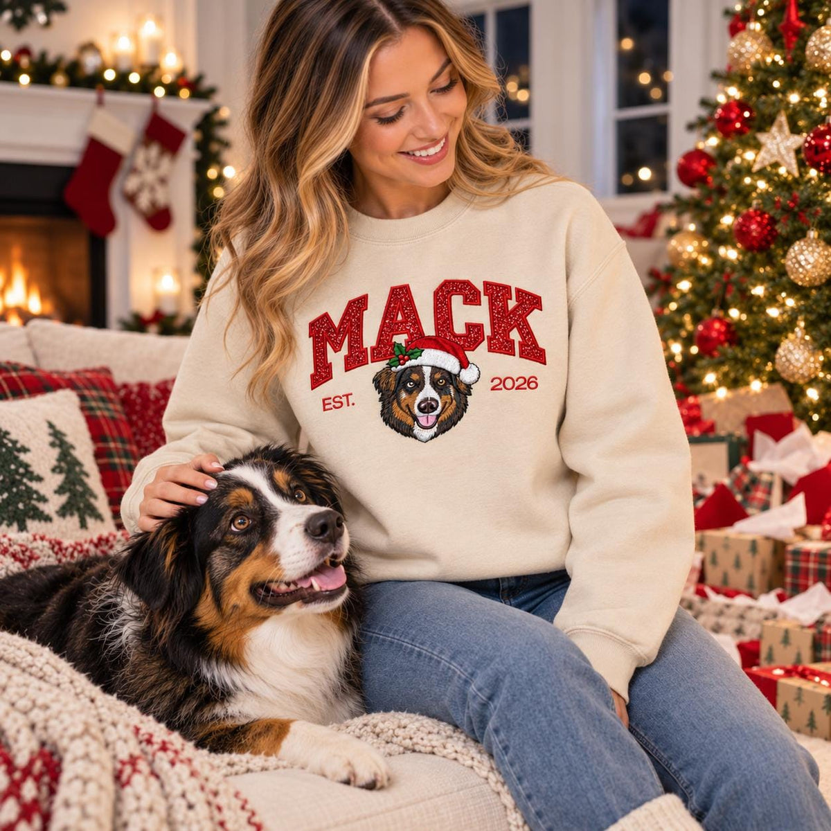 A smiling young woman wearing a custom Christmas dog sweatshirt in Sand color, featuring sparkly glitter text and a stitched Australian Shepherd wearing a Santa hat, petting her cute dog next to a glowing holiday tree.