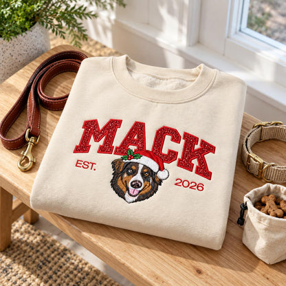 A flat lay product shot of a custom Christmas dog sweatshirt in Sand color placed on a wooden table, featuring sparkling red glitter embroidery, a festive Australian Shepherd portrait, a leather leash, and a treat bag.