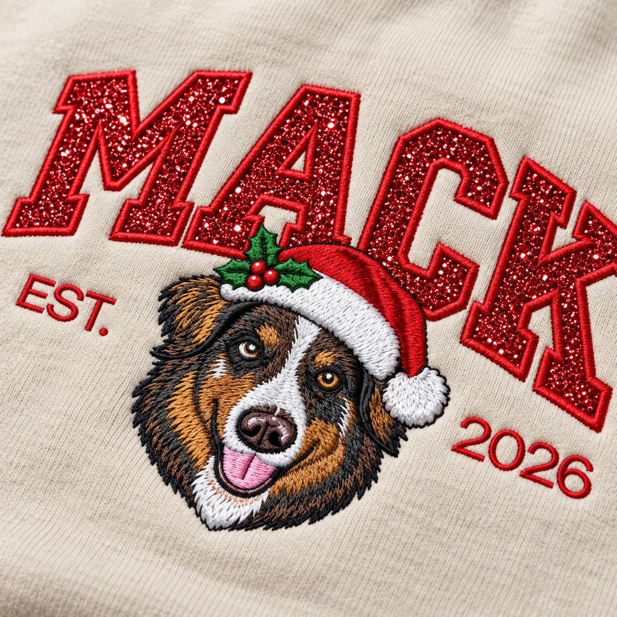 A macro close-up view of a Sand-colored custom Christmas dog sweatshirt, highlighting intricate red glitter letters and the vibrant embroidered face of an Australian Shepherd wearing a holiday Santa hat.