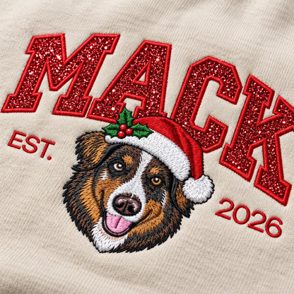 A macro close-up view of a Sand-colored custom Christmas dog sweatshirt, highlighting intricate red glitter letters and the vibrant embroidered face of an Australian Shepherd wearing a holiday Santa hat.
