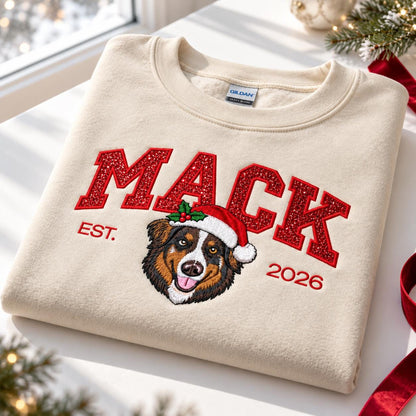 A folded Sand-colored custom Christmas dog sweatshirt resting on a white surface near a window, showcasing detailed red glitter embroidery, a festive Australian Shepherd wearing a Santa hat, and holiday decor.