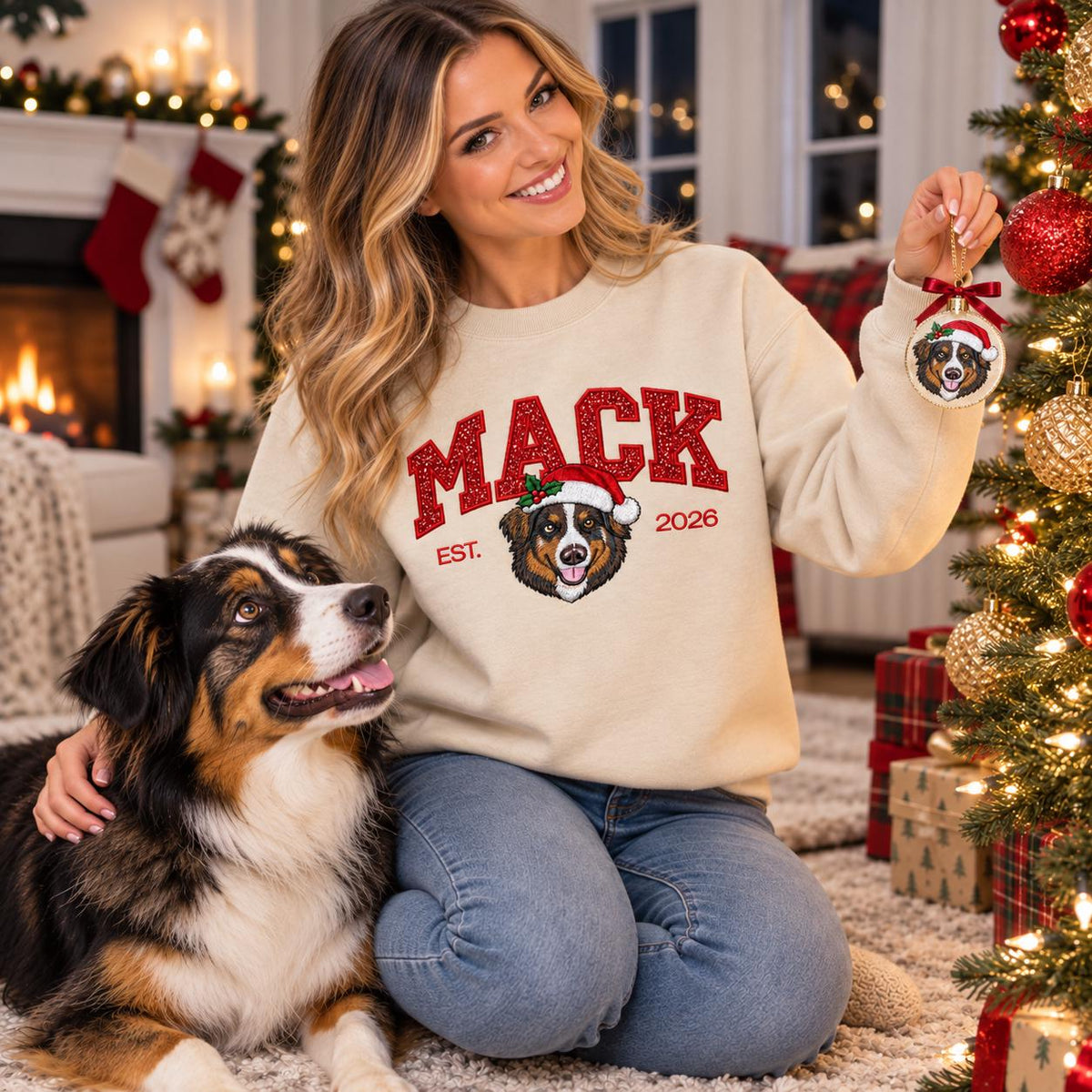 A woman wearing a Sand-colored custom Christmas dog sweatshirt holding a personalized holiday ornament, smiling warmly at her Australian Shepherd dog sitting happily by a beautifully lit Christmas tree.