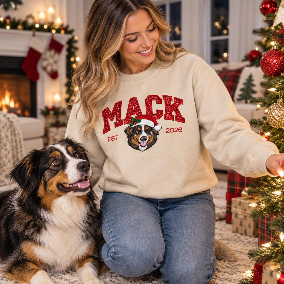 A woman smiling while sitting on a living room rug in a Sand-colored custom Christmas dog sweatshirt, admiring the twinkling lights of a decorated holiday tree next to her faithful Australian Shepherd dog.